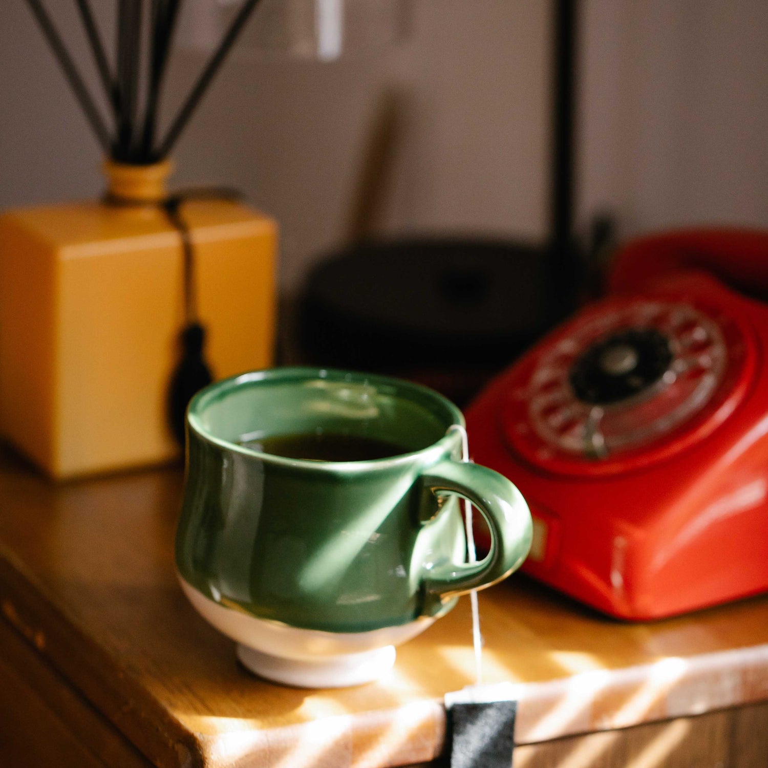 a cup of tea in a green mug with a reddish-orange rotary phone on a light wood table with a yellow reeded diffuser in the backbround