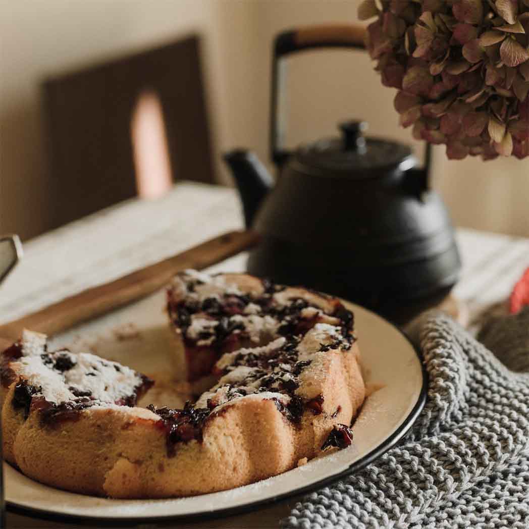 blueberry coffee cake on a plate sprinkled with powdered sugar.  black iron tea kettle in the background with dried hydrangeas, all on a table