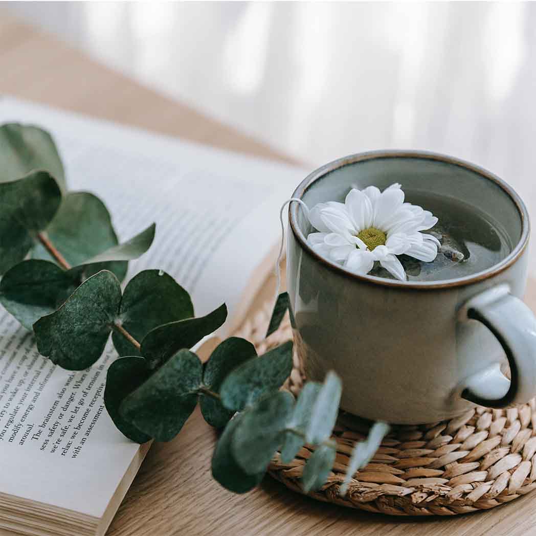 a cup of team sitting on a woven coaster next to an open book with a sprig of eucalyptus.  there is a white flower in the tea
