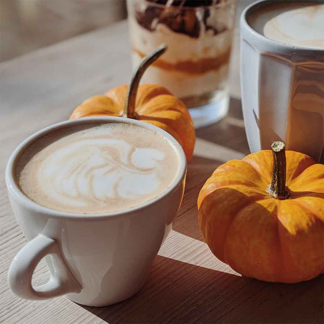 latte with foam art in a white mug on a table with two mini pumpkins and a caramel desert cup