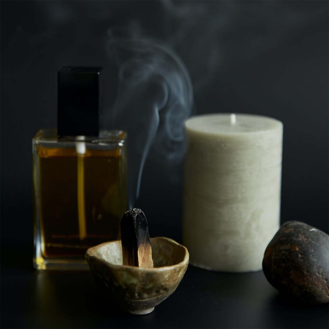smoking palo santo stick, a bottle of cologne, unlit off white pillar candle and a stone on a dark wood surface